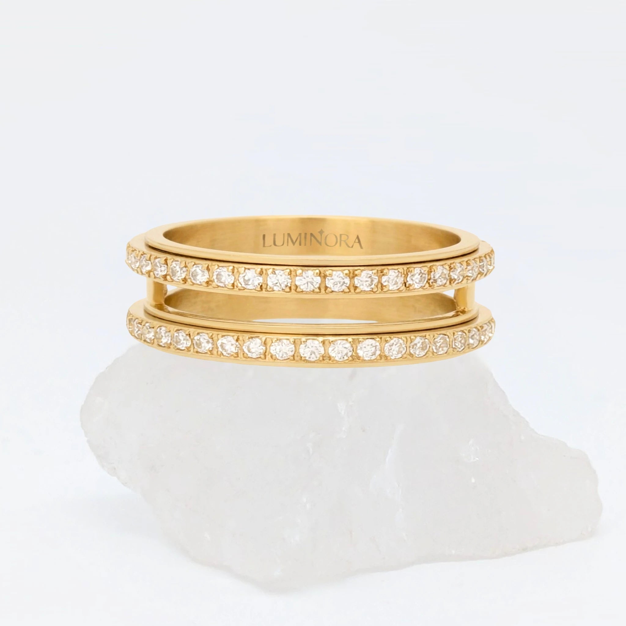Gouden Gemini fidget ring met een opvallende dubbele band vol fonkelende kristallen, gepresenteerd op een witte steen.