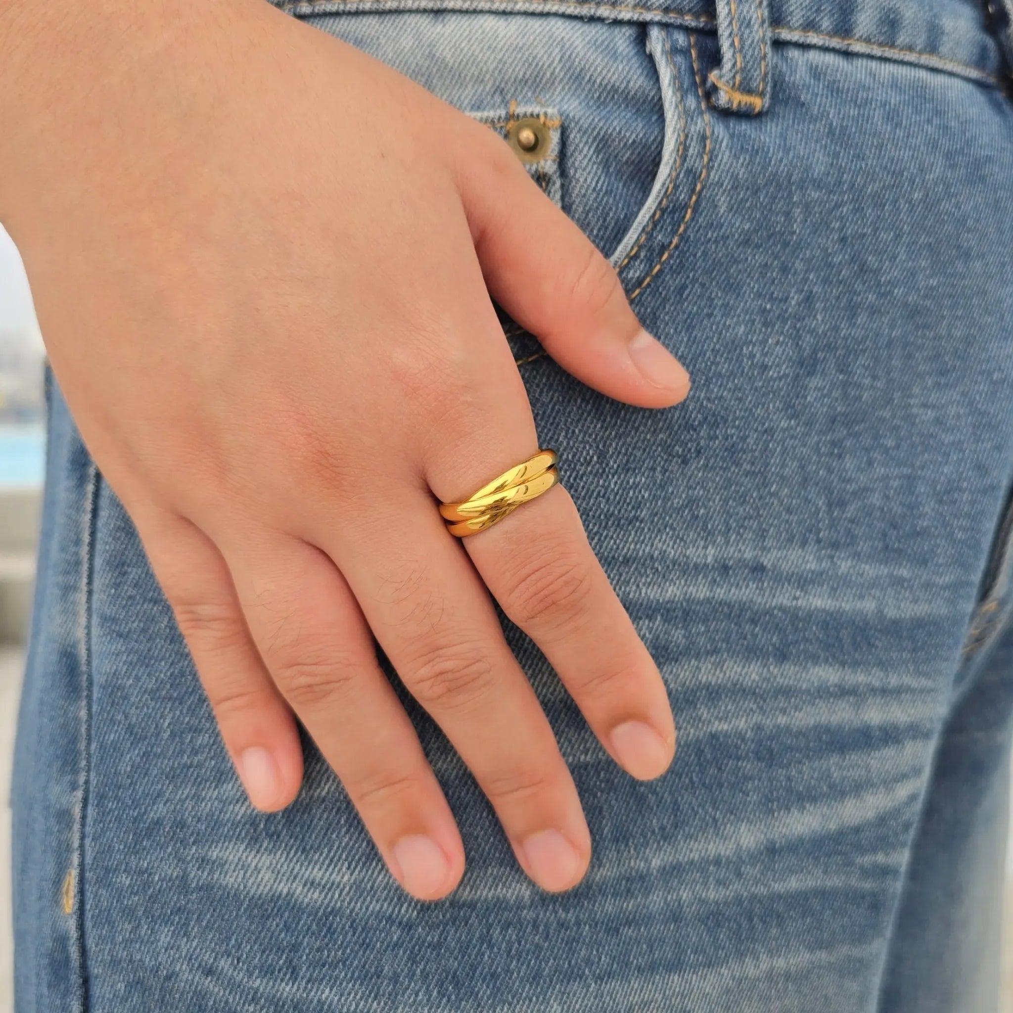 Hand van een vrouw met de gouden Trinity anxiety ring bestaande uit drie in elkaar grijpende RVS ringen, rustend op een blauwe spijkerbroek.