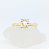 Gouden Halo fidget ring met een vierkant design en een parel in het midden, liggend op een witte steen.