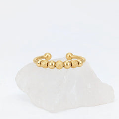 Gouden Ivy Bead fidget ring met een open design en afwisselend gladde en getextureerde balletjes, gepresenteerd op een witte steen.