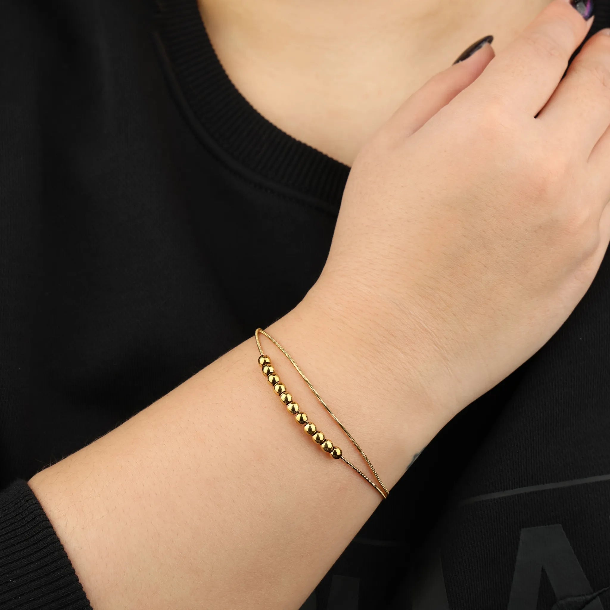 Vrouwenhand draagt goudkleurige Bead anxiety armband van RVS bij zwart shirt – Luminora