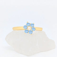 Gouden Aqua fidget ring in de vorm van een bloem met blauwe steentjes gepresenteerd op een witte steen.