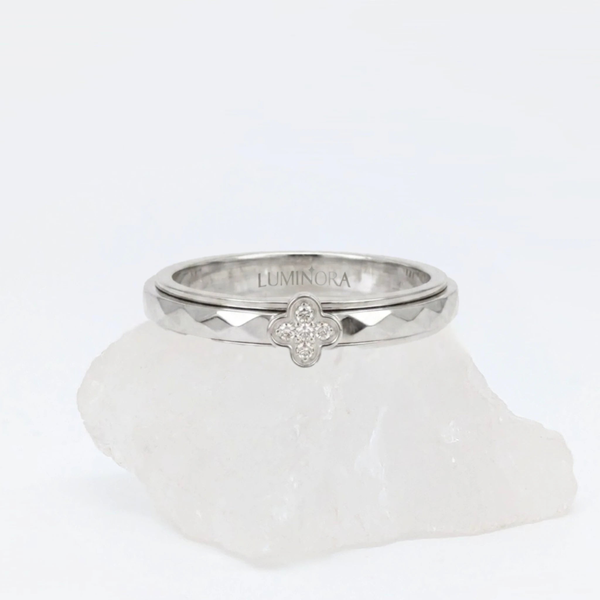 Zilveren Clover anxiety ring met een glanzend klavertje vier bezet met steentjes en een geometrisch gefacetteerde band, gepresenteerd op een witte steen.