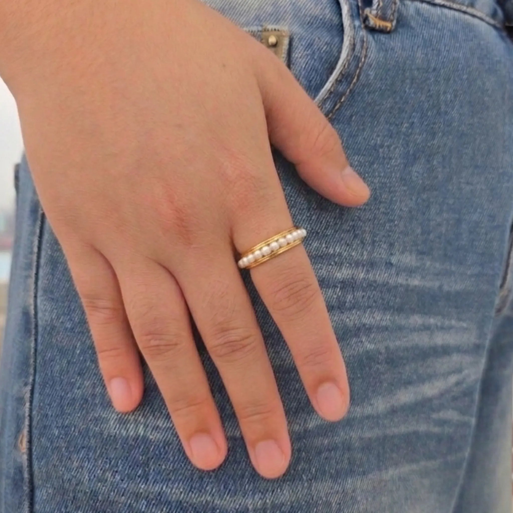 Hand van een vrouw met de gouden Perla anti-stress ring met parels om de vinger, gefotografeerd op een blauwe jeans.