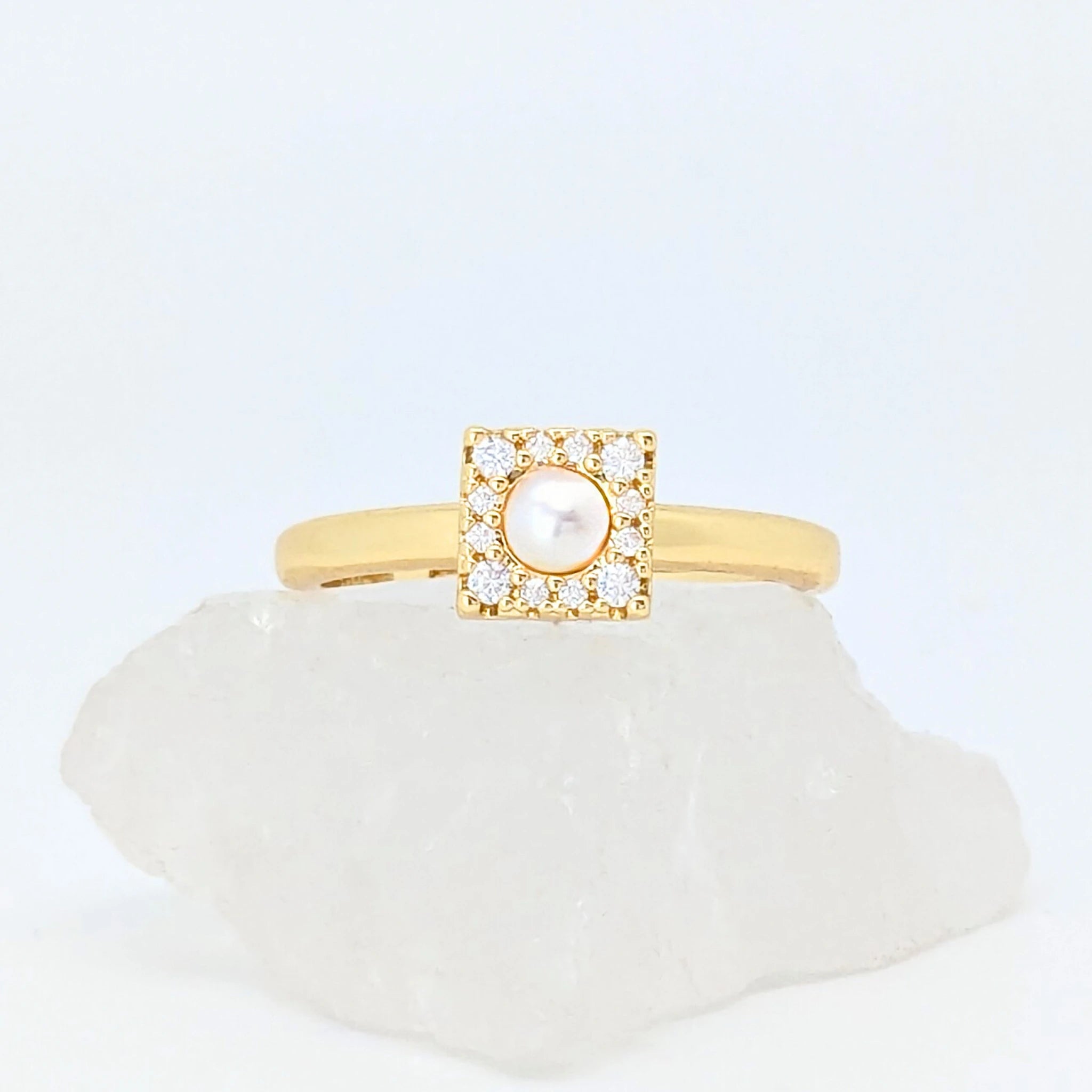 Gouden Halo fidget ring met een vierkant design en een parel in het midden, liggend op een witte steen.