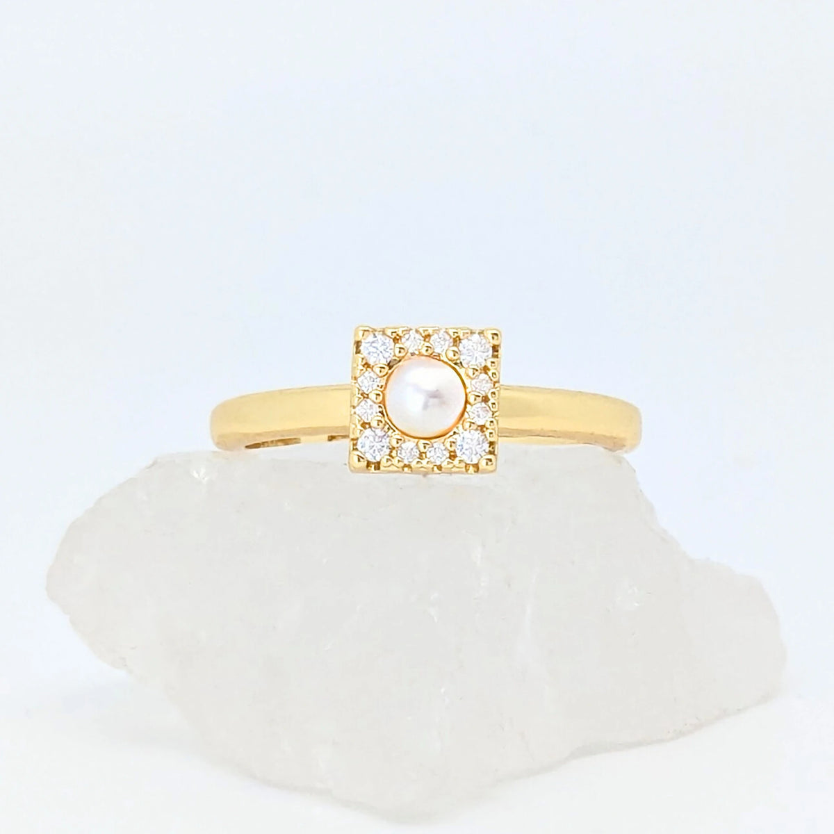 Gouden Halo fidget ring met een vierkant design en een parel in het midden, liggend op een witte steen.