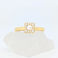 Gouden Halo fidget ring met een vierkant design en een parel in het midden, liggend op een witte steen.