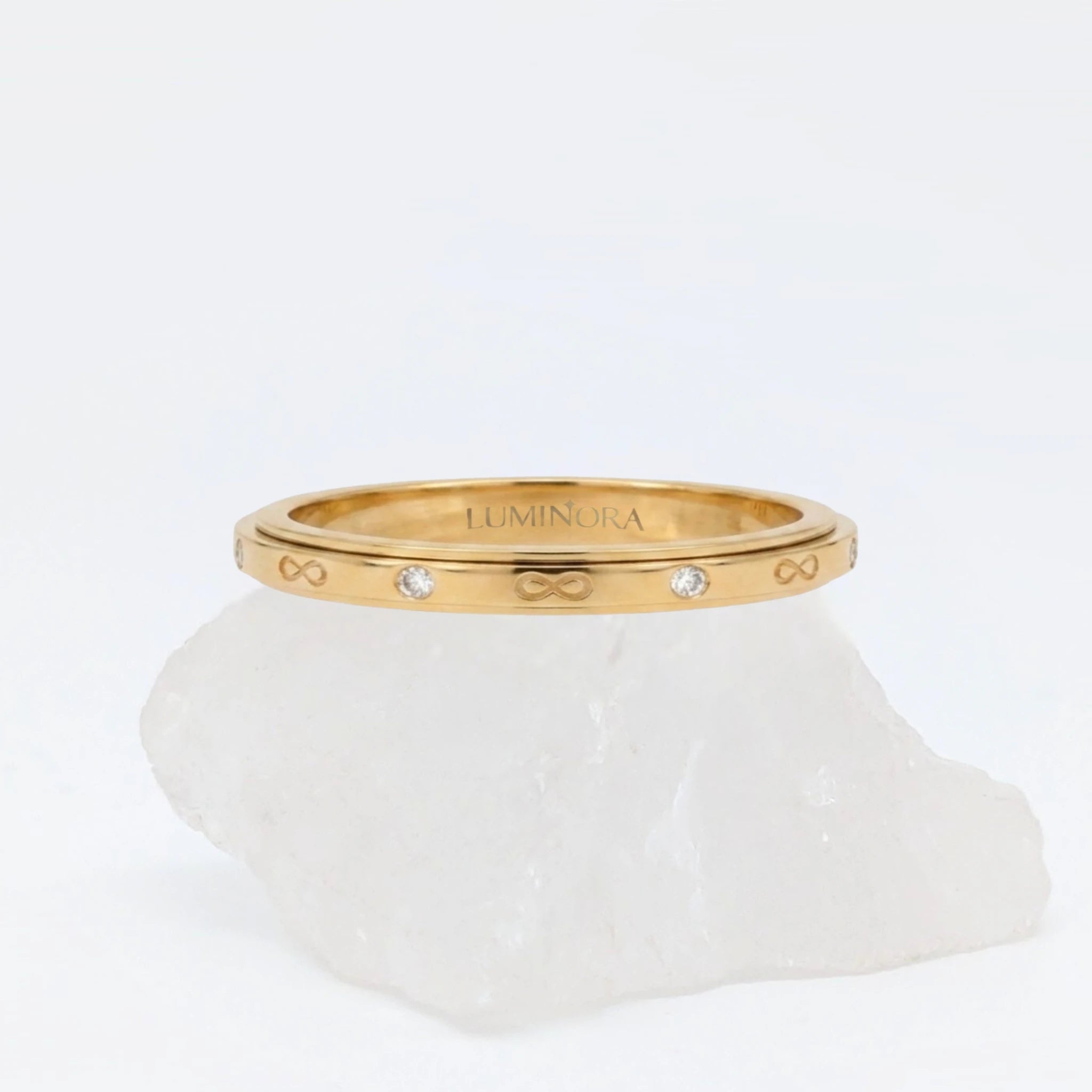 Gouden Infinity fidget ring met gegraveerde oneindigheidssymbolen en subtiele kristallen, gepresenteerd op een witte steen.