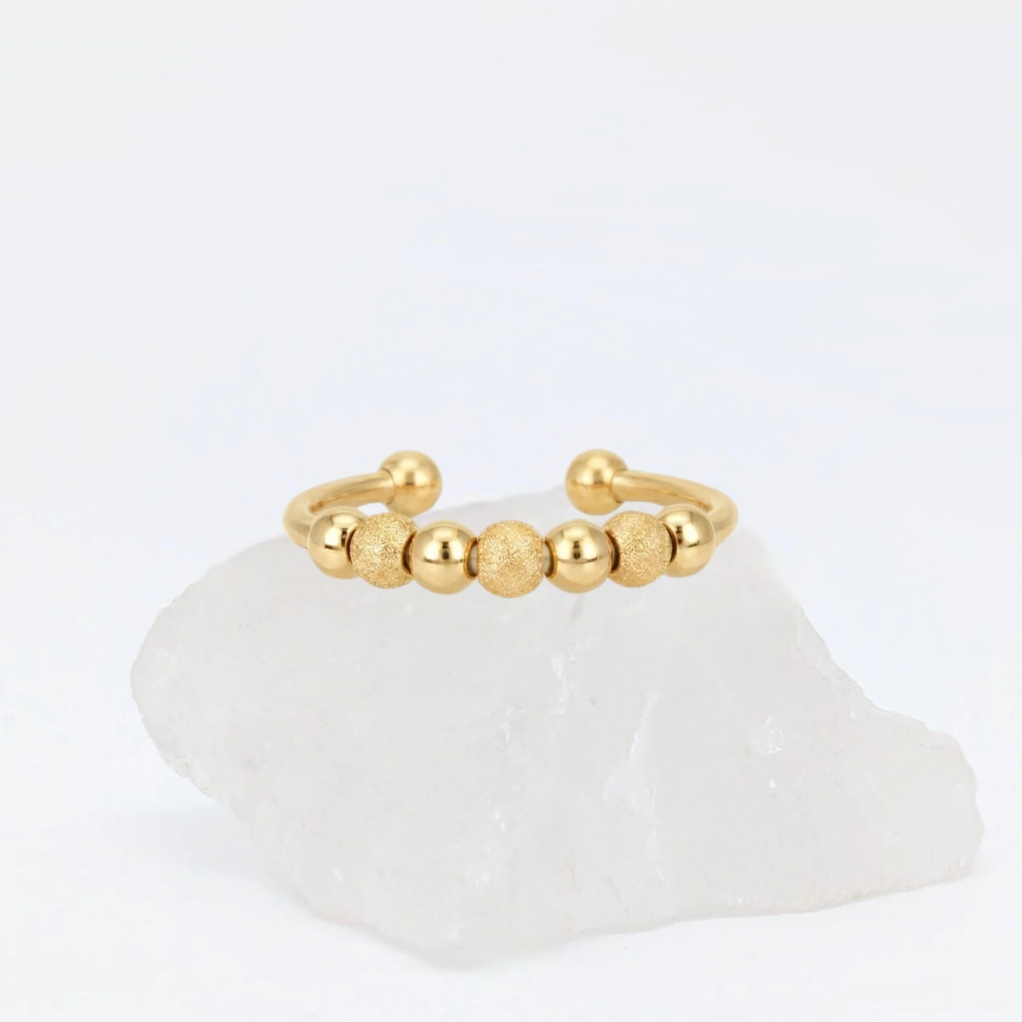 Gouden Ivy Bead fidget ring met een open design en afwisselend gladde en getextureerde balletjes, gepresenteerd op een witte steen.
